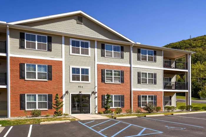 Attractive three-story apartment building with modern architecture, red brick facade, and inviting balconies set against a lush green backdrop.