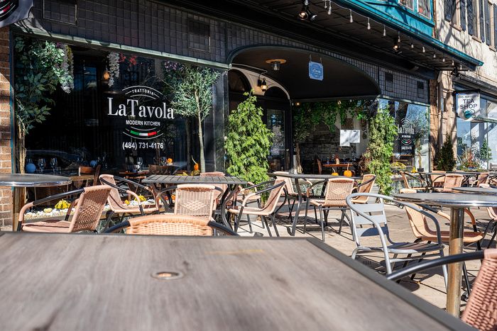 Charming outdoor dining area at La Tavola Modern Kitchen, surrounded by lush greenery and rustic wicker chairs.