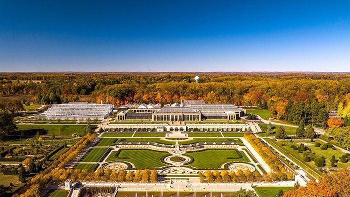 Breathtaking aerial view of a vast, elegant estate surrounded by lush autumnal forests, featuring manicured gardens and grand greenhouses under a clear blue sky.