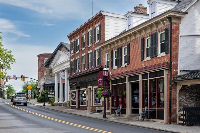 Charming downtown street view featuring historic brick buildings with large windows, cozy outdoor seating areas, and vibrant hanging flower baskets under a bright blue sky.
