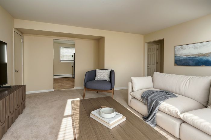 Cozy living room with plush beige sofa, modern armchair, and elegant decor, leading to a kitchen area with natural light.