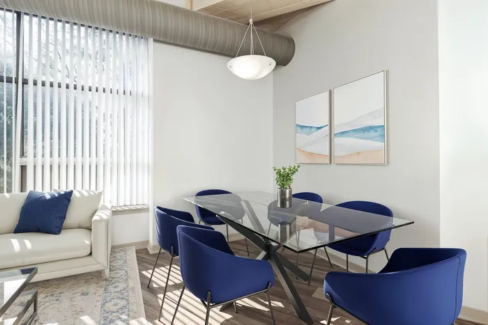 Bright and modern dining area featuring a sleek glass table, stylish blue chairs, and a cozy sofa with natural light streaming through vertical blinds.