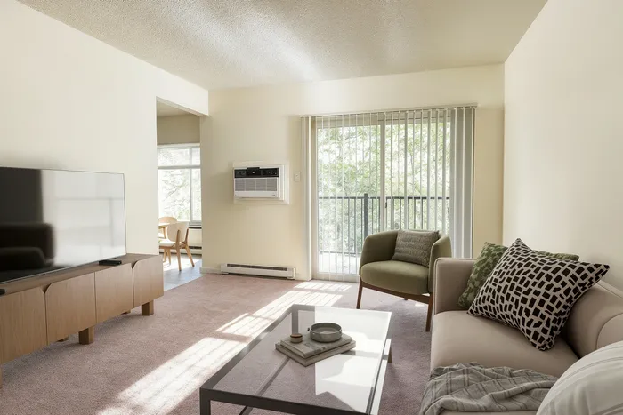 Bright living room with plush carpet, modern furniture, and a large TV unit, leading to a sunlit balcony with lush green views.