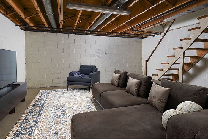 Chic basement lounge featuring a plush sectional sofa, cozy armchair, and stylish area rug under exposed beams and industrial lighting.