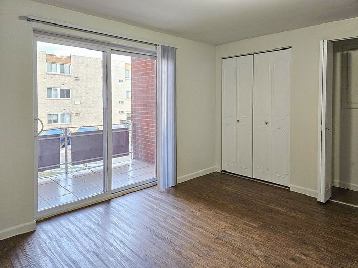 Bright apartment room with sleek hardwood floors, large sliding glass doors opening to a private balcony, and stylish white closet doors.