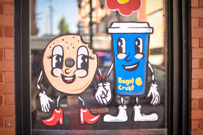 Charming window art featuring a cartoon bagel character with red boots and a smiling blue coffee cup labeled 'Bagel & Crust'.