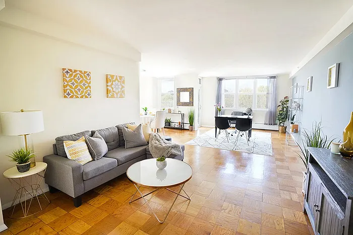 Bright and inviting living area with a modern gray sofa, elegantly paired with decorative cushions and a chic coffee table; sunlight floods through large windows, highlighting the spacious dining space and stylish decor.
