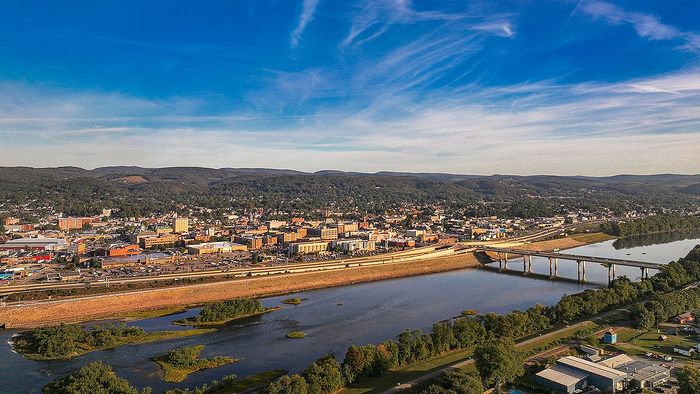 Scenic aerial view of a vibrant town surrounded by lush greenery, a tranquil river, and picturesque hills under a bright blue sky.