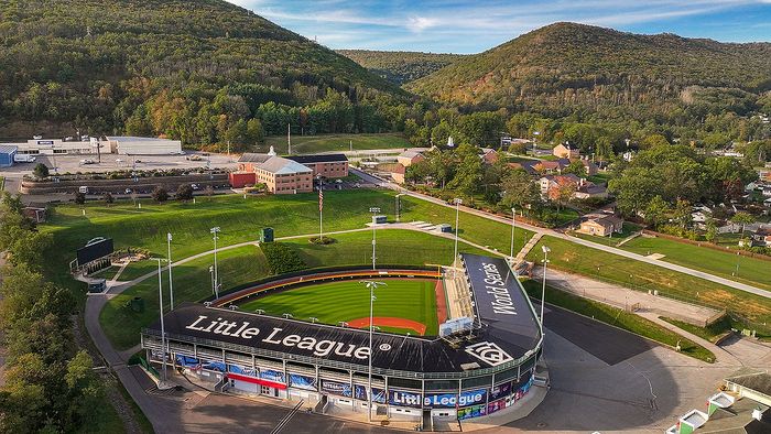 Scenic aerial view of a vibrant baseball stadium nestled among lush green hills, featuring the iconic Little League branding and surrounded by charming community buildings.