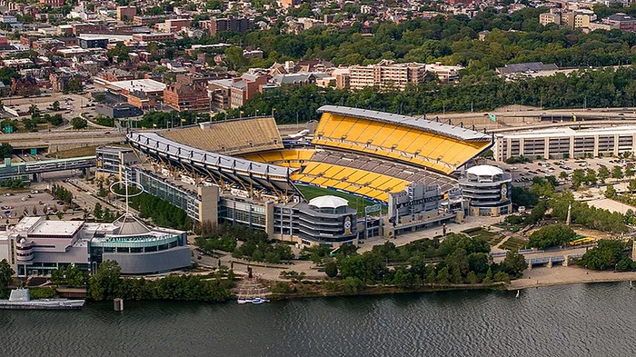Aerial view of a vibrant sports stadium with striking yellow seats, surrounded by lush greenery and a scenic river, set against a bustling urban backdrop.