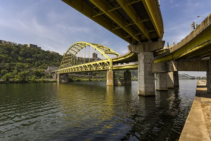 Vibrant yellow arch bridge spanning over a tranquil river, set against a lush green hillside and clear blue skies.
