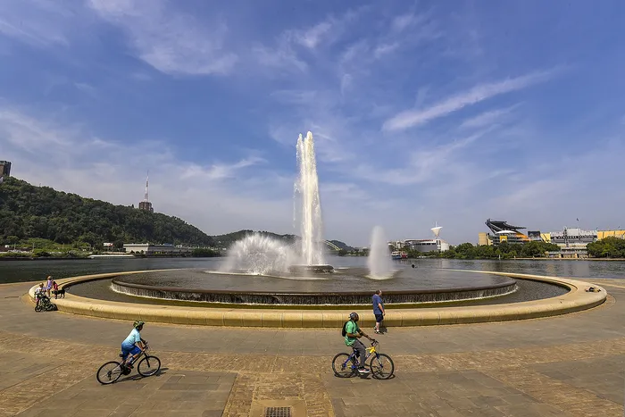 Vibrant cityscape with a stunning waterfront fountain, cyclists enjoying a sunny day, and scenic hills in the background.