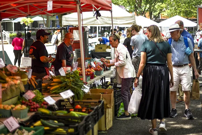 Vibrant farmers market scene with fresh produce and cheerful vendors under red canopies, engaging with happy customers.