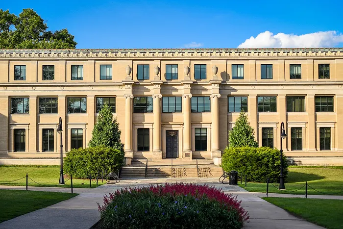 Elegant neoclassical building facade with grand columns and lush garden setting, bathed in sunlight beneath a clear blue sky.