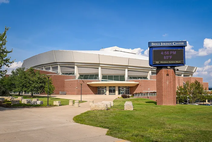 Modern architectural stadium with a sleek design, large entrance, and a digital marquee displaying time and temperature, set against a bright blue sky.