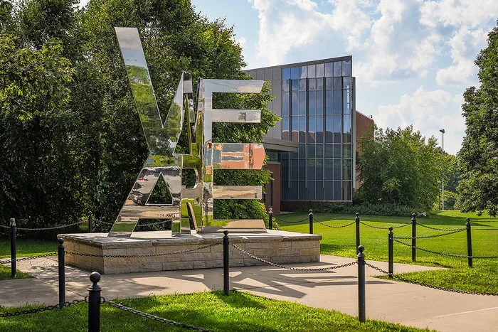 Modern glass-fronted building with lush green surroundings and a contemporary mirrored sculpture.