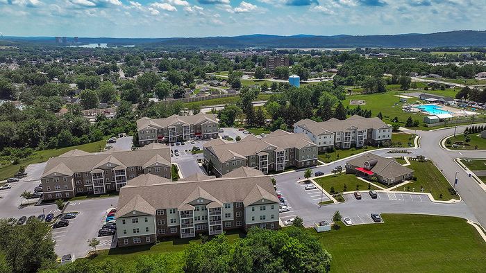 Aerial view of a modern apartment complex surrounded by lush greenery, featuring spacious parking and a refreshing pool area, ideal for comfortable living.