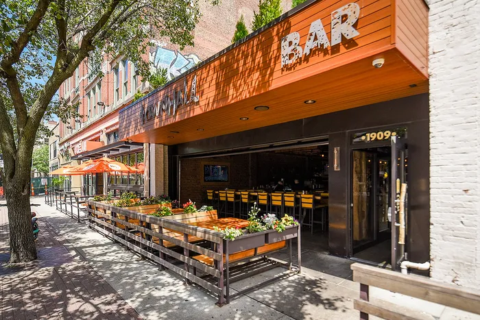 Vibrant urban scene featuring a stylish bar with a welcoming outdoor patio, adorned with colorful flower beds and orange umbrellas.