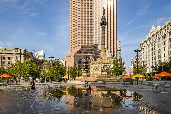 Vibrant urban plaza with a striking fountain, historic monument, and towering city buildings under a clear blue sky.