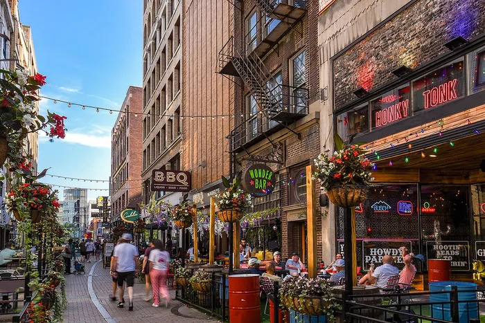 Vibrant city alley with lively restaurants and hanging flower baskets under a clear blue sky.