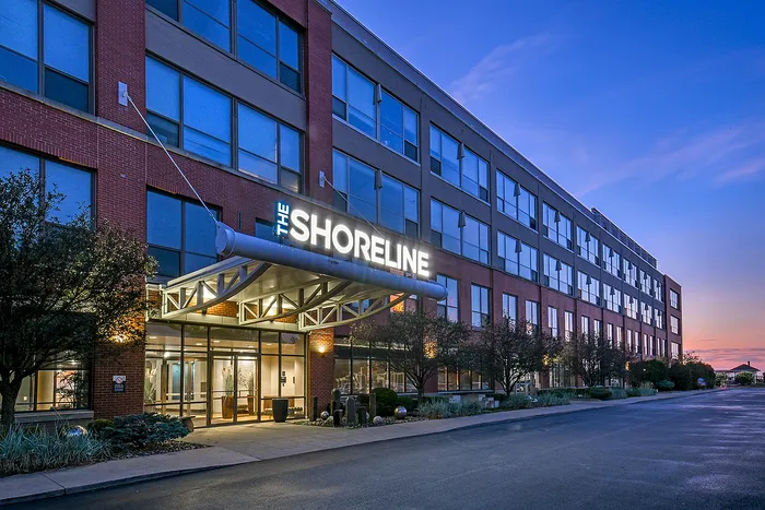 Elegant exterior of The Shoreline apartments at dusk, showcasing modern architecture with illuminated signage and expansive glass windows.