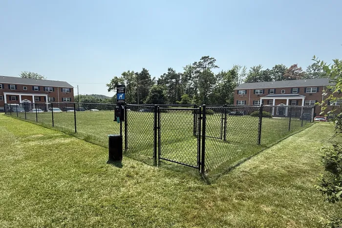 Charming fenced dog park surrounded by lush green grass in front of brick apartment buildings, with clear blue skies above.