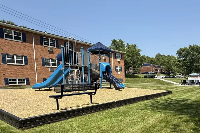 Inviting playground with vibrant blue slides, nestled in a spacious grassy area adjacent to charming brick apartment buildings.
