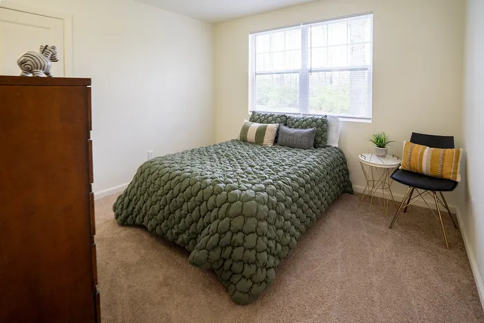 Inviting bedroom with cozy green quilted bedding, bright natural light from large windows, and stylish accent chair and table.