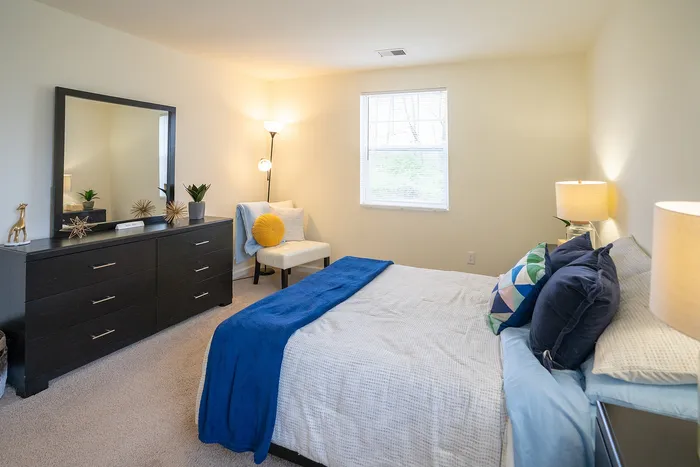 Inviting bedroom with stylish decor, featuring a comfortable bed adorned with vibrant pillows, a sleek black dresser with decorative accents, and a cozy seating area by a bright window.