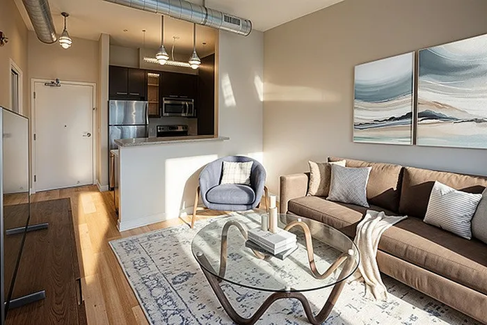 Modern living room featuring a sleek brown sofa, elegant glass coffee table, cozy armchair, and stylish kitchen with stainless steel appliances.