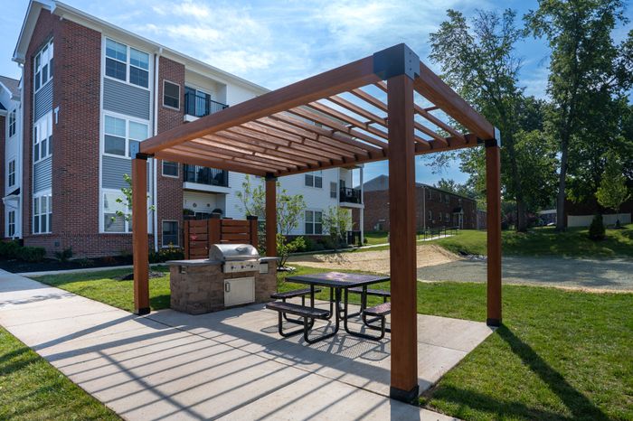Inviting outdoor area with a modern wooden pergola, featuring a stylish barbecue grill and seating, set against a backdrop of a charming brick apartment building and lush greenery.