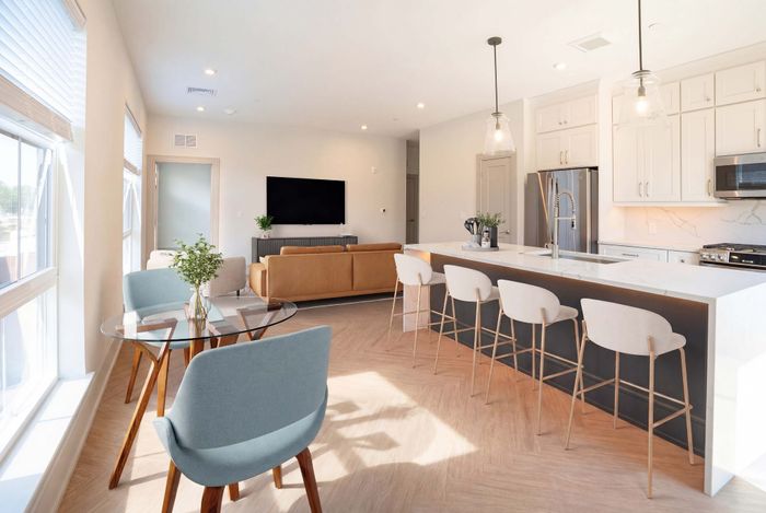 Bright modern living space with a stylish kitchen island, comfortable seating area, and a glass dining table next to large windows.
