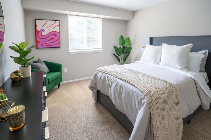 Inviting bedroom with cozy gray upholstered bed, plush pillows, and vibrant artwork above a green accent chair, bathed in natural light.