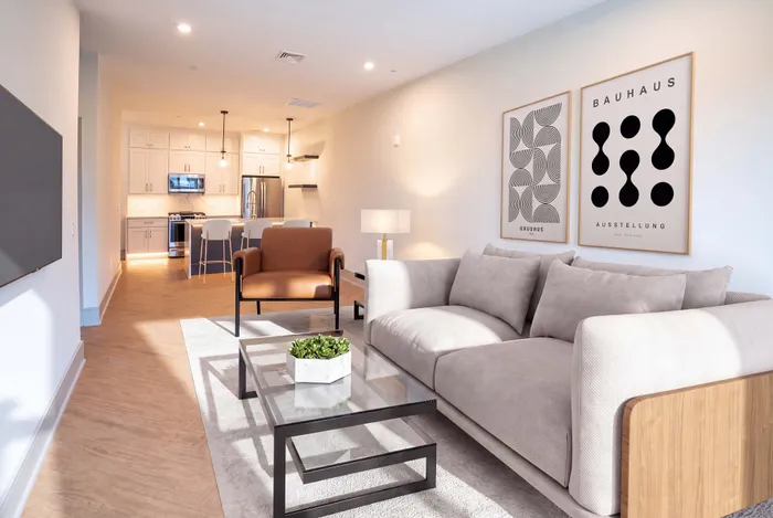 Inviting modern living room with cozy gray sofa, stylish armchair, and sleek glass coffee table adjoining a contemporary kitchen with chic lighting and bar seating.