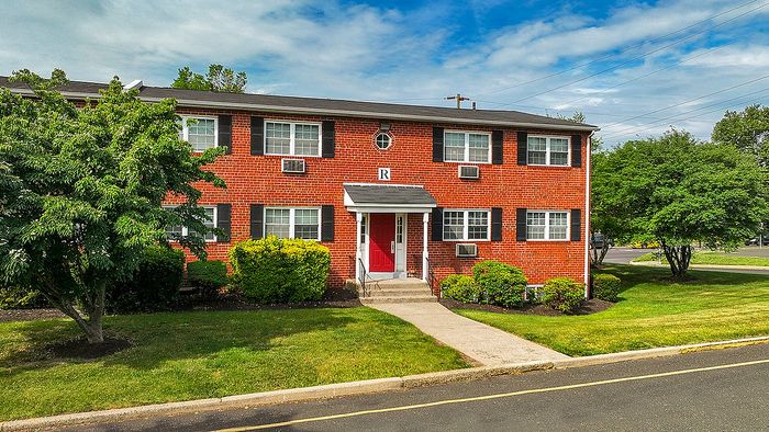 Charming brick apartment building with lush greenery and a welcoming red door, set against a bright blue sky.