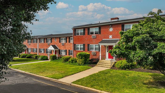 Charming brick apartment complex with lush green landscaping and inviting red doors, perfect for a serene living experience.