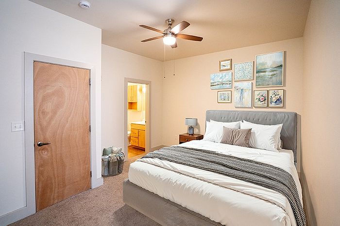 Inviting bedroom with plush bedding, elegant wall art, and cozy carpet, featuring an en-suite bathroom and modern ceiling fan.