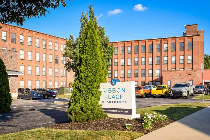 Charming Ribbon Place Apartments with classic red-brick exterior surrounded by lush greenery, ample parking space, and a welcoming entrance sign.