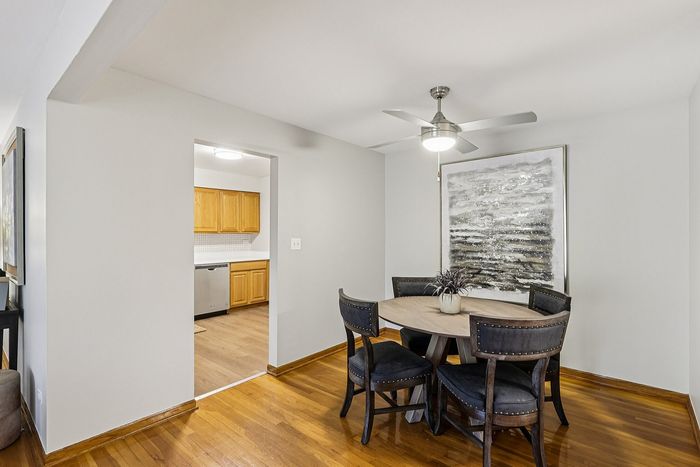 Chic dining area with elegant round table and modern black chairs, complemented by a stylish ceiling fan and abstract wall art, adjacent to a bright kitchen with warm wood cabinets.