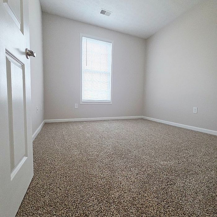 Bright and inviting empty room with plush carpet flooring and a large sunlit window, perfect for creating your ideal living space.
