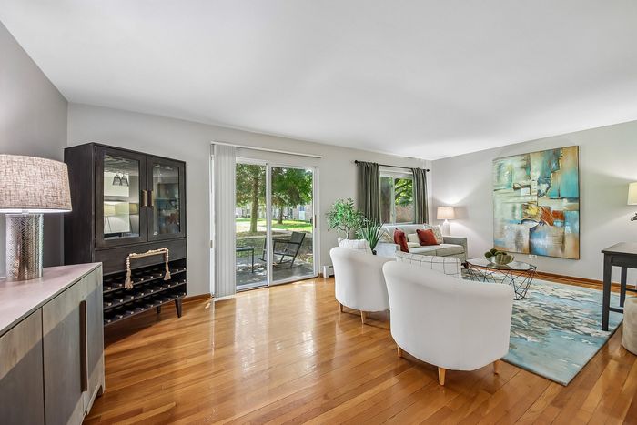 Elegant living room with hardwood floors, stylish white chairs, and modern art, offering abundant natural light through large sliding doors.