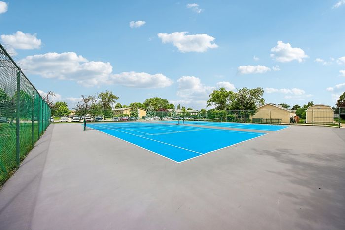 Vibrant blue tennis court surrounded by lush greenery and residential houses under a clear blue sky.