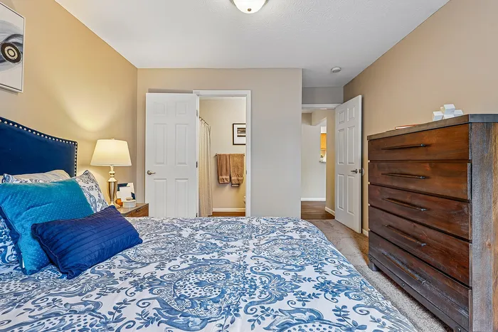 Inviting bedroom with elegant decor, featuring a plush blue and white bedspread, wooden dresser, and a soft-lit lamp creating a cozy ambiance.