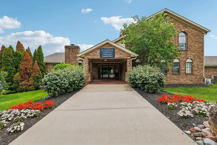 Inviting entrance of WoodSpring Apartments with lush landscaping and colorful flower beds under a bright blue sky.