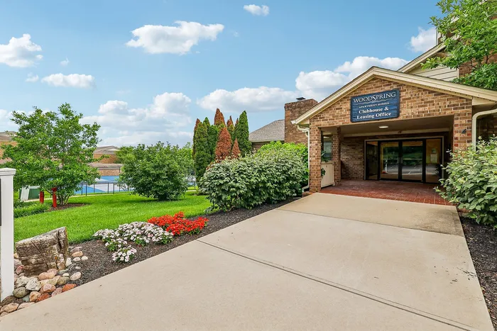 Inviting entrance to Woodspring apartment homes, featuring lush landscaping and modern brick architecture under a vibrant blue sky.