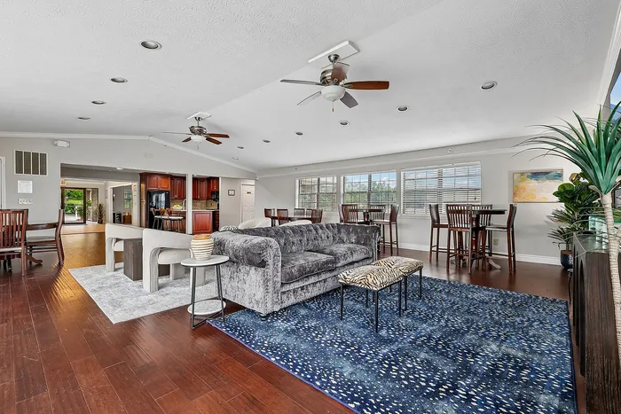 Expansive open-concept living space with elegant hardwood floors, modern ceiling fans, and ample natural light. Features a plush gray sofa, stylish dining area, and a vibrant blue area rug.