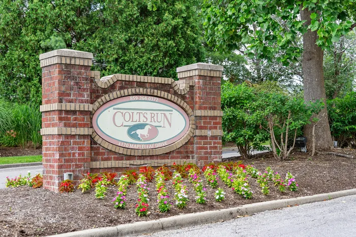 Inviting entrance to Colts Run with rustic brick signage and vibrant landscaped flowers against a lush green backdrop.