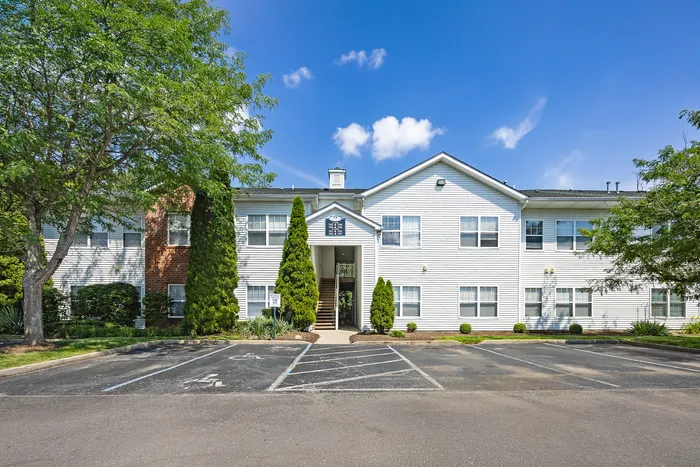 Charming apartment building surrounded by lush greenery, featuring a welcoming entrance and ample parking space under a vibrant blue sky.