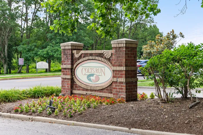 Inviting entrance to Colts Run with elegant brick signage surrounded by lush greenery and colorful flower beds.