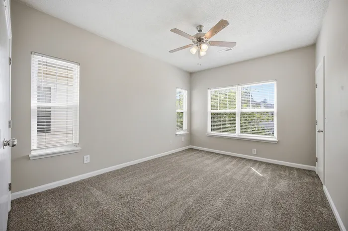Bright and spacious bedroom featuring large windows with natural light, modern ceiling fan, and plush carpeting.